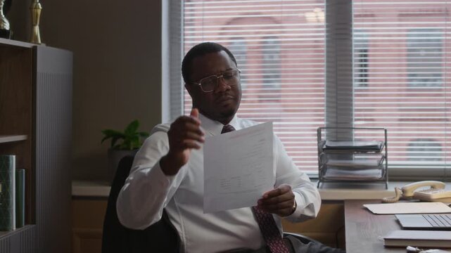 Side view of young black male principal in formalwear sitting at desk while printing documents in school office