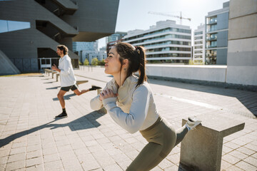 A woman smiles and stands next to a man as they hold their hands together in front of them and lean their feet on stone chairs while doing lunges