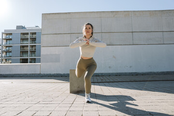 A woman lunges with her feet resting on stone chair and her hands together in front of her