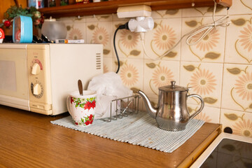 Cozy retro kitchen corner with an old microwave, festive poinsettia mug and spoon, and a shiny metal teapot resting on a mat against patterned flower tiles.