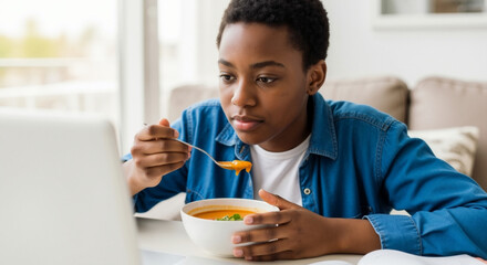 Black teen boy eating healthy vegetable soup while looking at laptop screen. African American student having lunch break at home