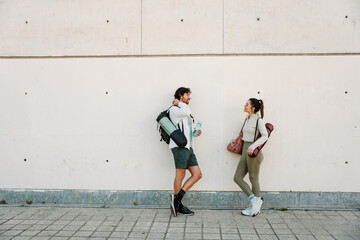 A man and a woman stand and lean on a wall while holding a mat and smiling while he talks and holds a bottle