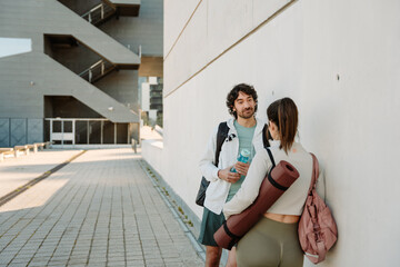 A man holds a bottle and listens in surprise to a woman standing across from him as they lean on a wall