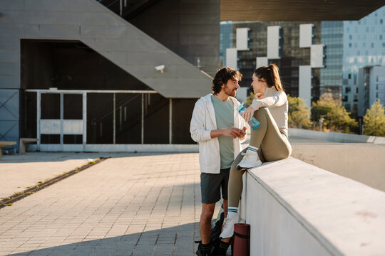 A man stands and talks to a woman who is holding a bottle and sitting on a wall while smiling - Powered by Adobe
