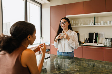 A female worker stands and smiles while holding a cup and listening to a female worker sitting across from her at a table