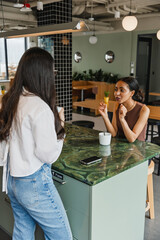 A female worker sits on a chair at a table and talks to a female worker who is holding a cup and...