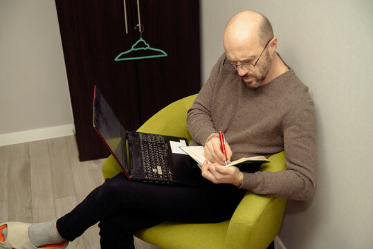 Focused man working remotely at home, writing notes in a notebook while using his laptop. Ideal for themes of freelance work, online learning, productivity, and comfortable home office environments.