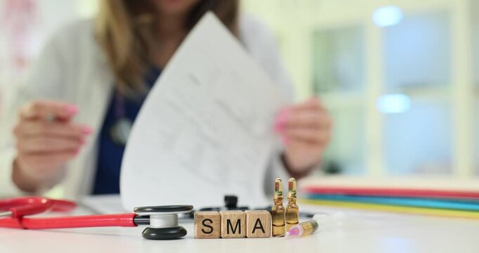 Miniature wooden cubes on table form abbreviation SMA near ampoules and stethoscope. Woman doctor reviews notes in notebook with focused attention