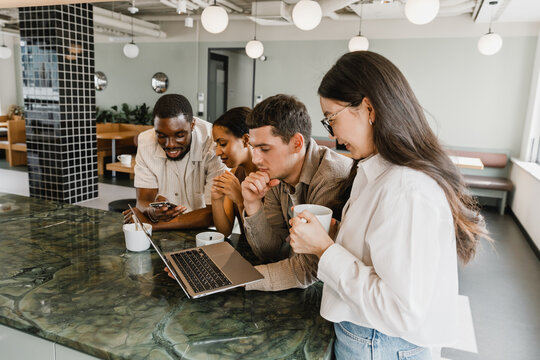 Group of four workers at a table in pairs looking at a laptop and phone while talking and smiling
