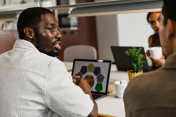 Male worker in front of laptop and listening to male worker next to him while they sit at table