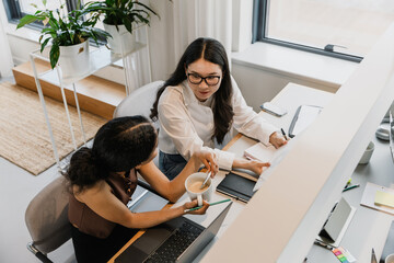 Female worker holding a folder and talking to a female worker holding a cup while they sit at a table