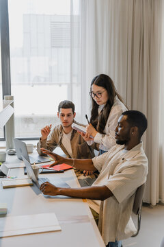 A group of three workers are looking at a laptop while two of them are sitting at a desk and one of them is standing and holding a notebook