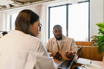 Obraz premium A male worker points at a laptop he is looking at with a female worker across from him who is talking while they are sitting at a table