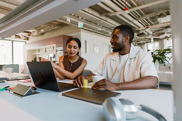 Man holding stickers and smiling while looking at female worker looking at laptop while they are sitting at table