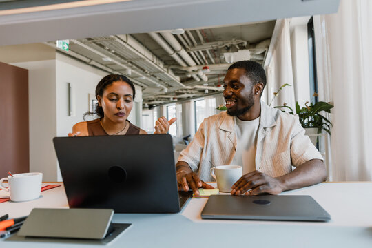 Male worker laughing and listening to female worker gesturing and looking at laptop while they sit at table