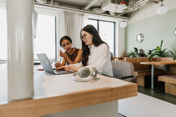 A female employee is talking and looking at a laptop while a female employee sitting next to her at the desk is scrolling through it