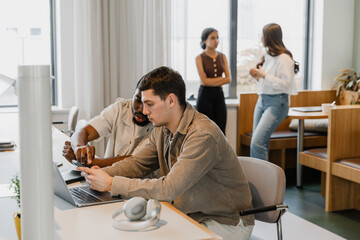 A male employee holds a phone, which a male employee is pointing at while they are talking while sitting at a desk