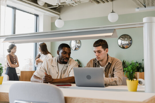 A male worker looks at a laptop in front of which a male worker sits while they are at a table