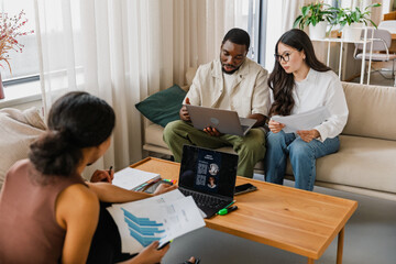 Male and female workers sitting on a couch looking at a laptop while he talks and she holds documents while two workers are in front of them