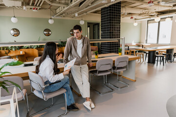 A male employee stands and leans on a table while talking to a female employee who is sitting on a chair and holding a document