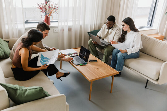 A group of four workers sit on sofas in pairs at a table and hold documents while looking at laptops