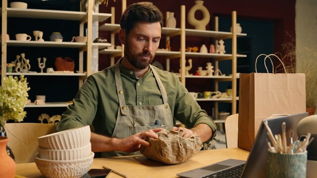 Caucasian man guy male small business owner crock businessman potter craftsman packing ceramic pottery bowl package porcelain order delivery safety product shopping online check laptop indoor workshop