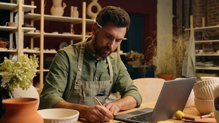 Caucasian salesman seller potter male man guy businessman artist small business owner examining bowl clay ceramic writing notes working new design laptop computer device inside pottery workshop store
