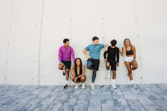 A group of five athletes are leaning on a wall and laughing while one of them is sitting and four of them are standing with their hands on each other's shoulders