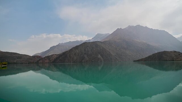A pan left view across turquoise water at Iskanderkul Lake in the Fann Mountains of Tajikistan, with rocky peaks reflected in a calm alpine landscape of Central Asia.