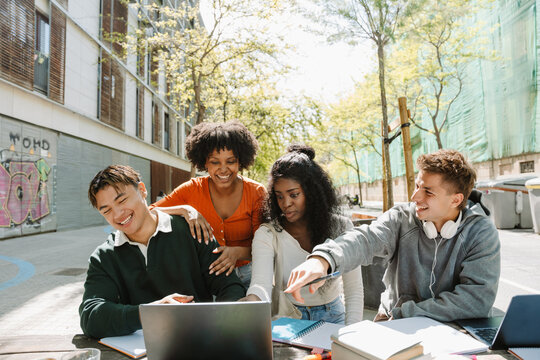 A group of four students are laughing and looking at a laptop on a table, which one of them is pointing at