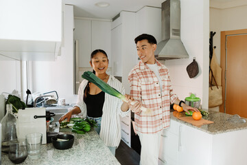 Man and woman holding green onions and smiling while standing at tables and she talking