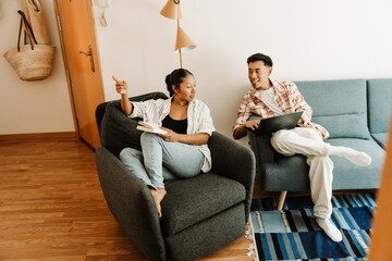 A woman is holding a book and sitting in a chair while talking to a man who is holding a laptop and sitting on a couch
