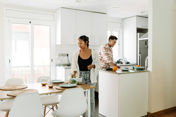 A woman puts a bowl of salad on the table while a man stands behind her and smiles