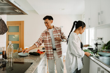 A man smiles and places a plate on the table while a woman stands next to him holding greens