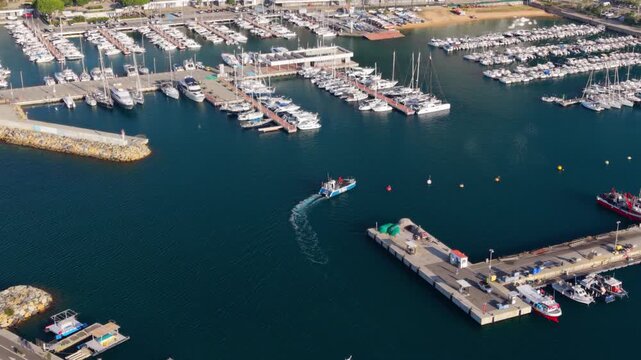 Majestic aerial view of the port of blanes in costa brava, spain, where a fishing boat is sailing out to sea while many yachts and sailboats are moored at the marina on a sunny summer day