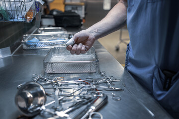 A healthcare professional in scrubs organizes surgical instruments on a stainless-steel worktable in a hospital sterilization or reprocessing department.