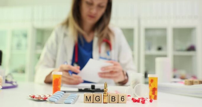 Woman doctor sits at desk and carefully writes prescription for vitamins. Wooden blocks forming abbreviation Mg B6 neatly arranged on office table