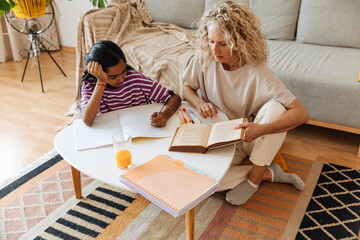 Daughter holds pen over notebook and looks at book her mother is reading while they sit at table
