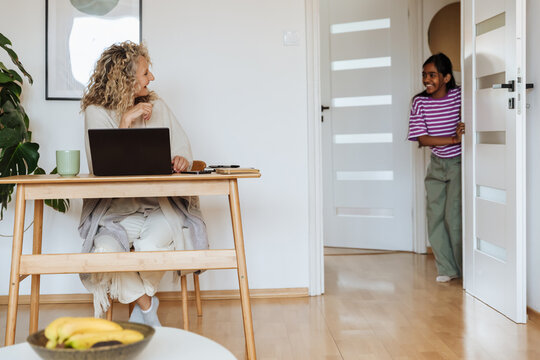 A mother sits on a chair at a table while her daughter looks out the door and they laugh