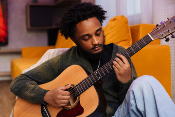 A male musician sits and plays the guitar, looking at it while leaning on a sofa