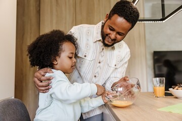 A father stands and smiles while hugging his daughter and holding a bowl in which she is stirring...