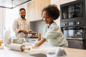 A father carries a baking dish and looks at his daughter who is standing at the table and spreading flour on the table