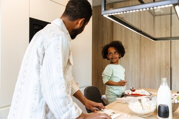Obraz premium A daughter stands on a chair at the table and holds a spoon over a baking dish while listening to her father standing next to her