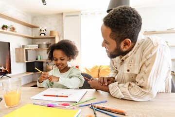 Daughter smiles and sits at the table while her father talks next to her while they hold pencils