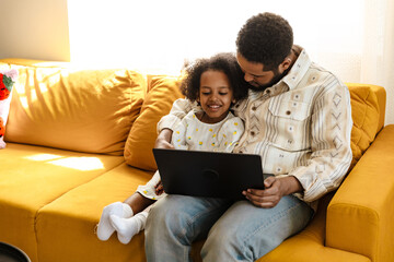 Fototapeta premium Daughter laughs and looks at laptop held by her father who hugs her while they sit on sofa