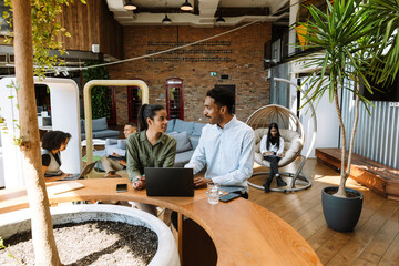 Male and female employees talking and smiling while sitting at a table in front of a laptop and looking at each other