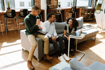 A group of three employees are laughing and sitting on a couch while one of them shows documents