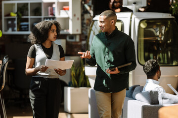 A male worker smiles and holds a computer tablet while talking to a female worker who walks next to...
