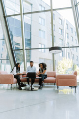 A group of three workers are looking at a laptop while two of them are smiling and sitting on a couch while the other is sitting on a chair