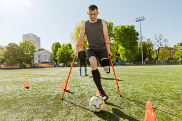A male player dribbles a ball between cones and leans on crutches while another male player stands behind him